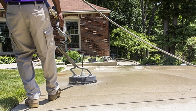 A stainless steel pressure washer surface cleaner being used on a sidewalk, with a bush and lawn in the background.