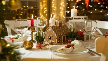 A table beautifully decorated with holiday candles, lanterns, and a gingerbread house as the centrepiece on a white tablecloth.