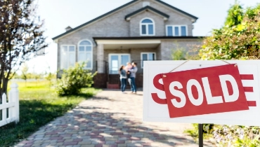 Sold sign in front of a home with young couple and baby blurred in the background.