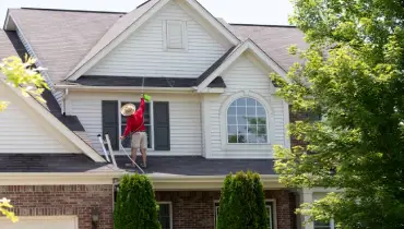 Homeowner washing the exterior of his house standing on the lower roof to spray the second-floor facade with a handheld pressure sprayer.